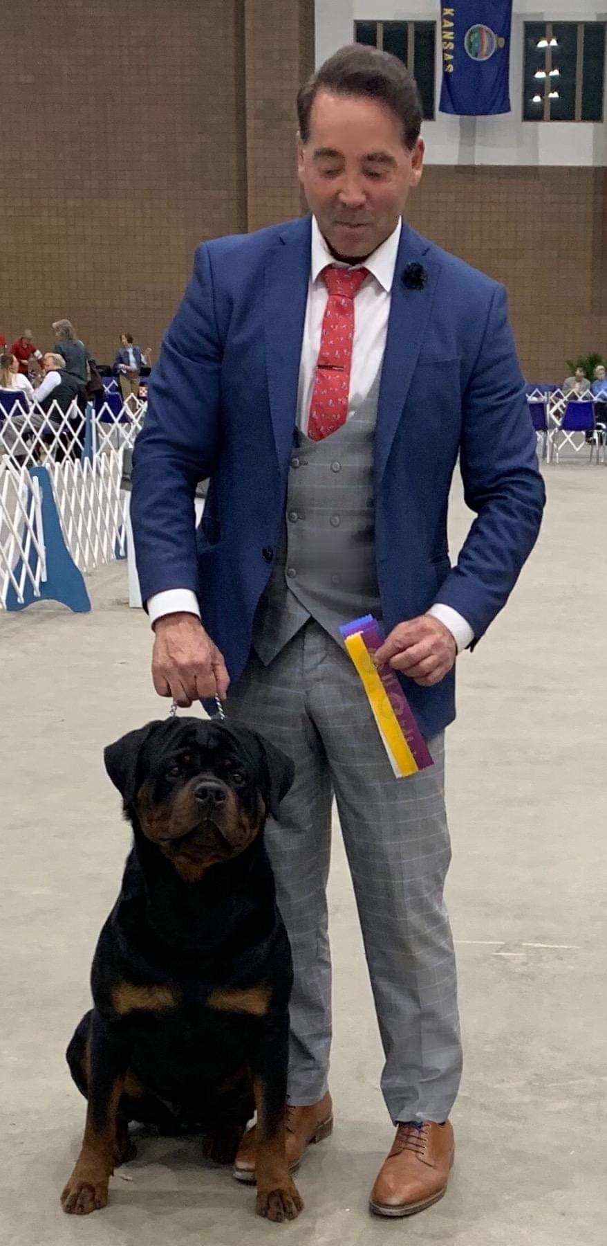 Arlos sitting at his handler's side in an indoor show ring, the handler in a blue suit holding a ribbon, a Kansas flag on the wall behind