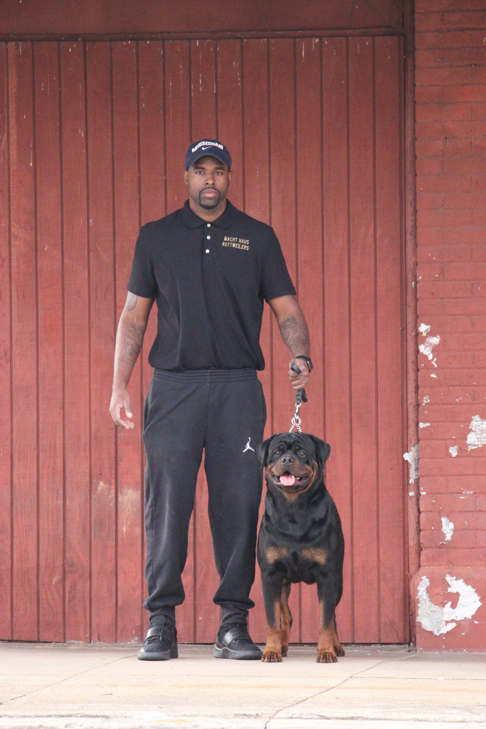 Arlos standing on lead beside his owner, who wears a Macht Haus Rottweilers polo, against a weathered red board wall