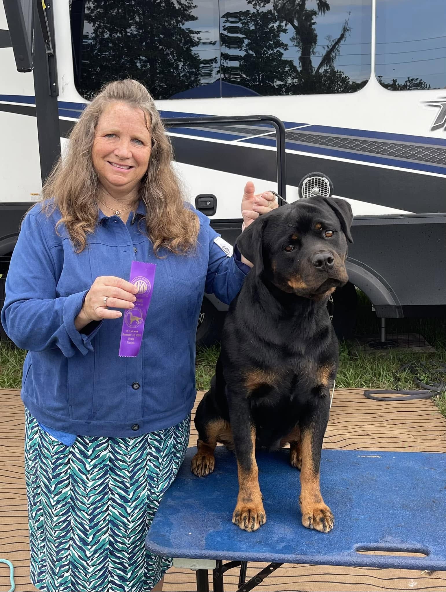 Journey standing on a grooming table beside her handler at a show, the handler holding a purple ribbon, an RV behind them