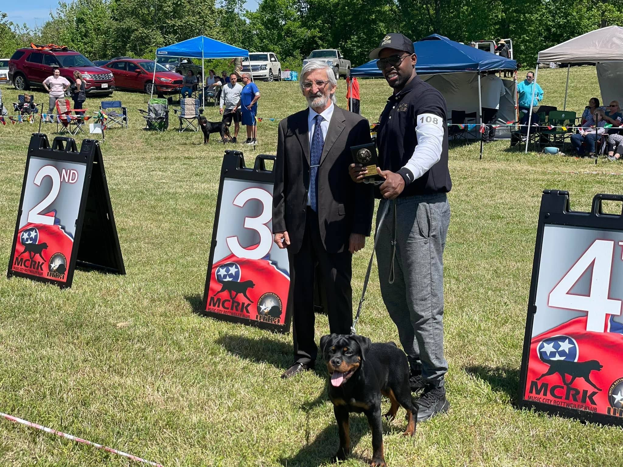 Truth standing on lead between her handler and the judge at a Music City Rottweiler Klub show, the handler holding a placement trophy