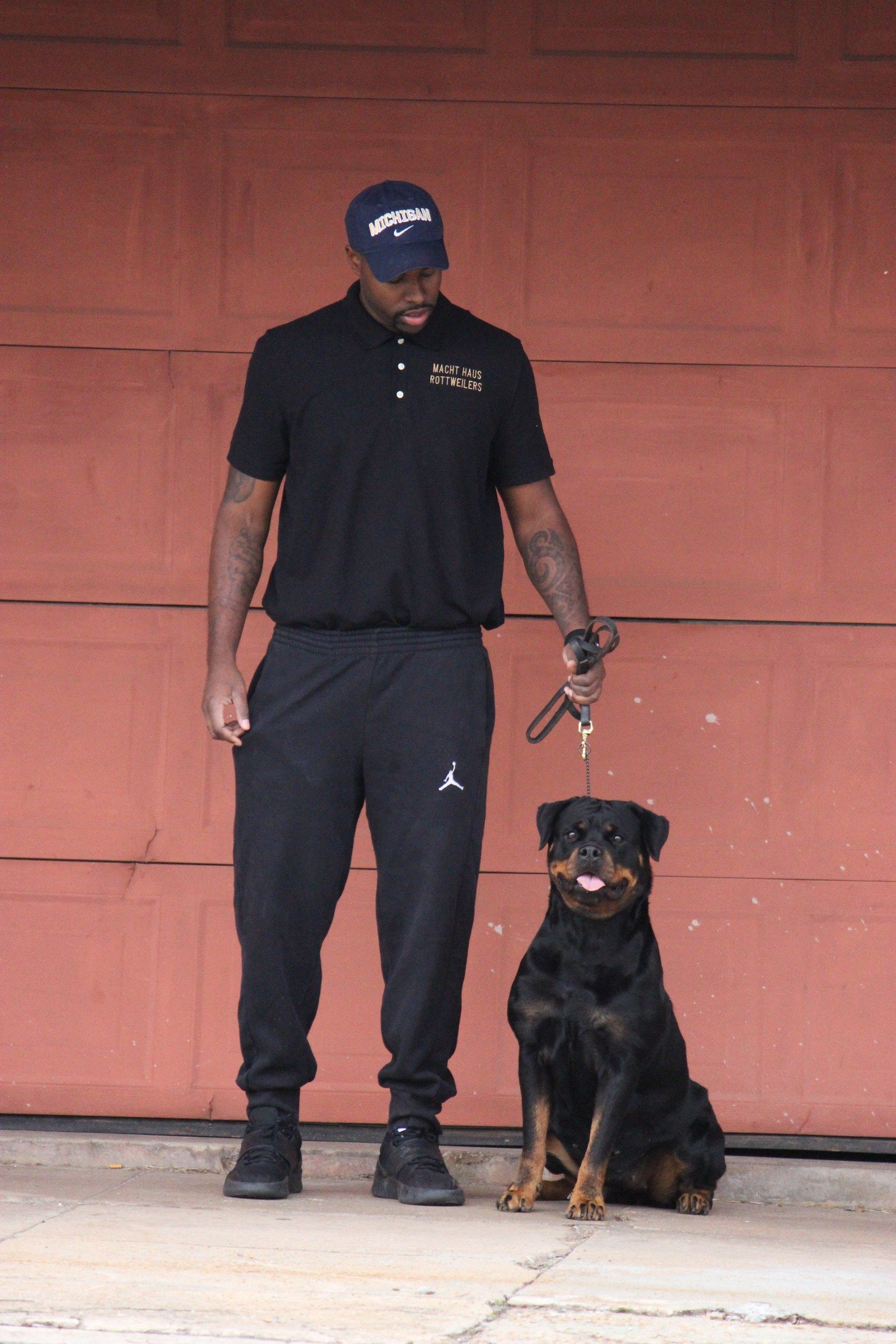Hera sitting beside her handler against a red garage door, the handler looking down at her