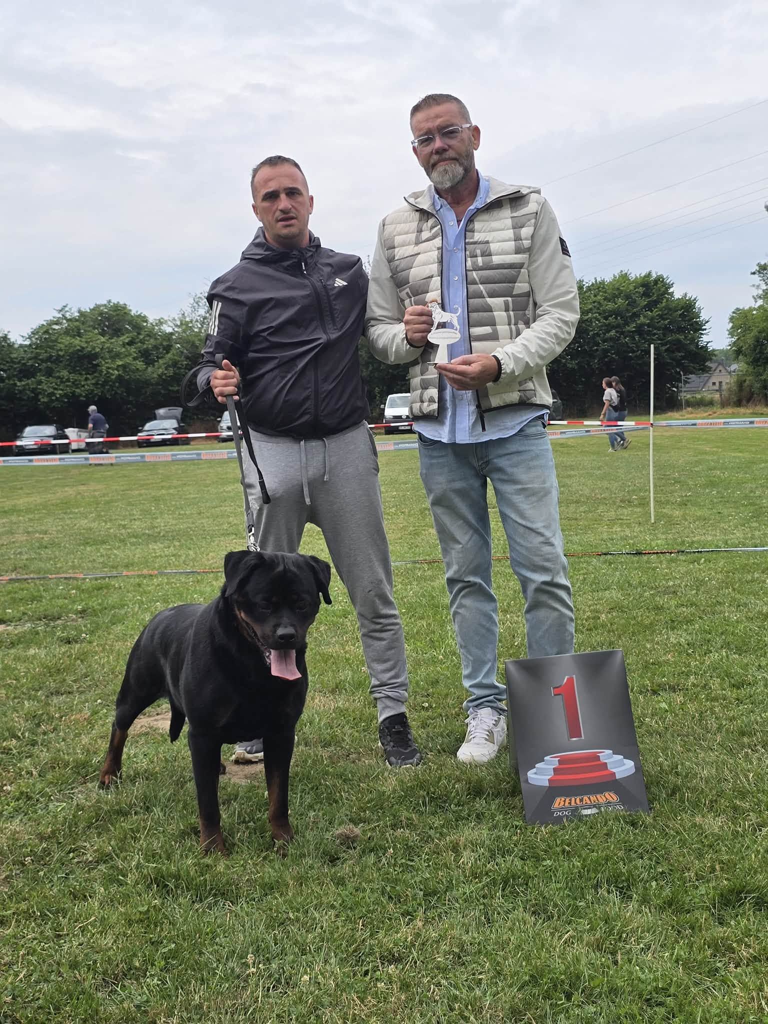 Trixi standing on lead beside her handler and the judge at an outdoor European show, a first-place sign and small trophy beside them